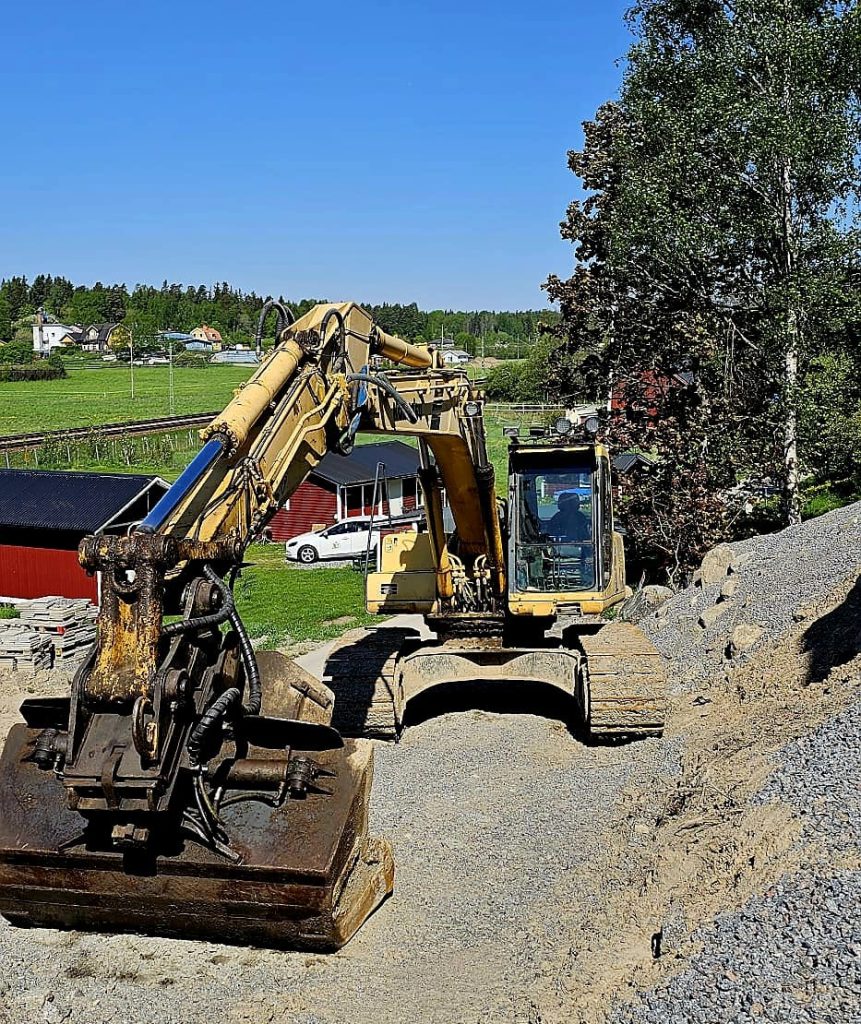 Grävmaskin på grusplan med fält och träd i bakgrunden.